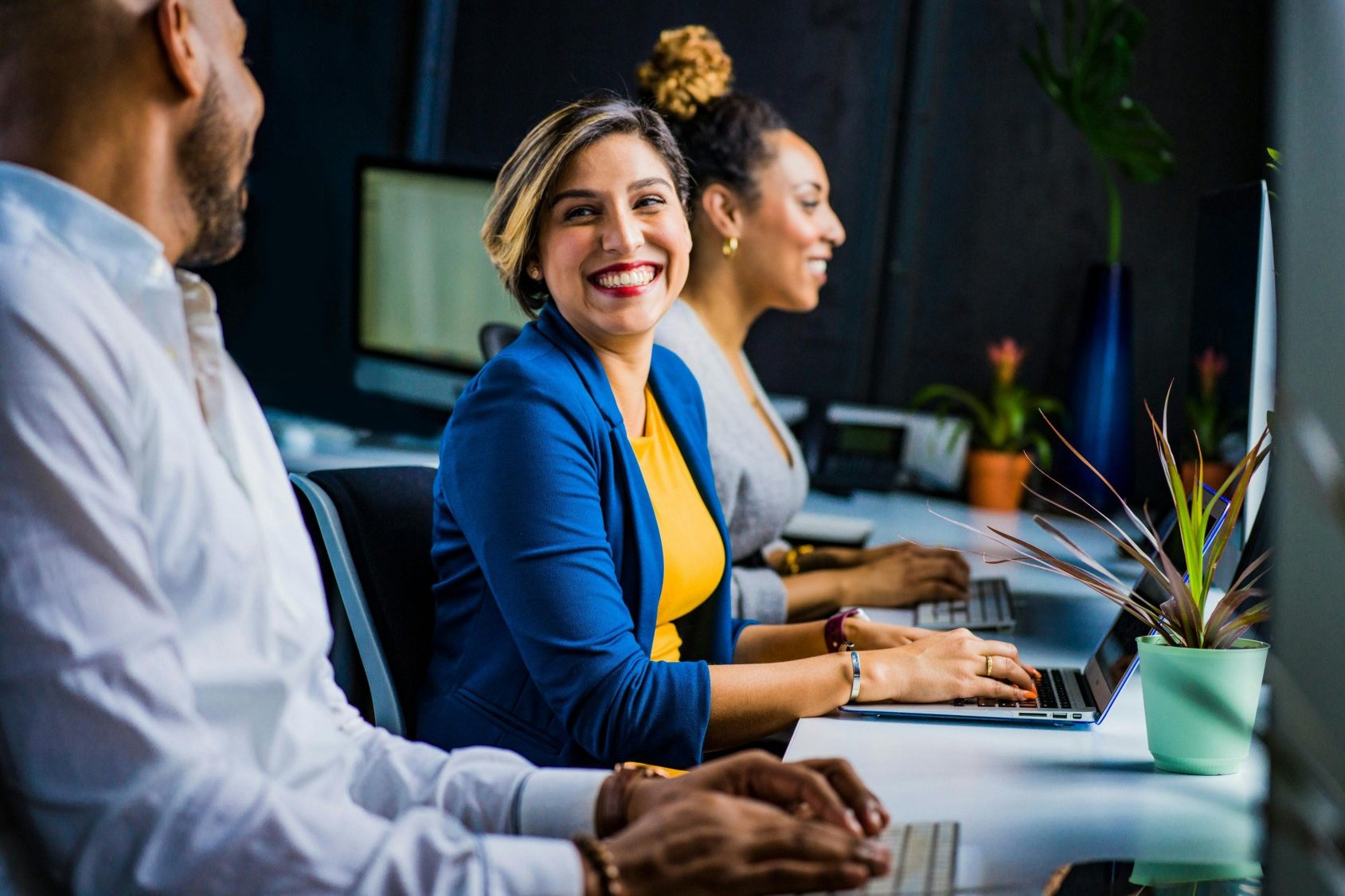 Three diverse professionals working and smiling at office desks, fostering teamwork and collaboration.